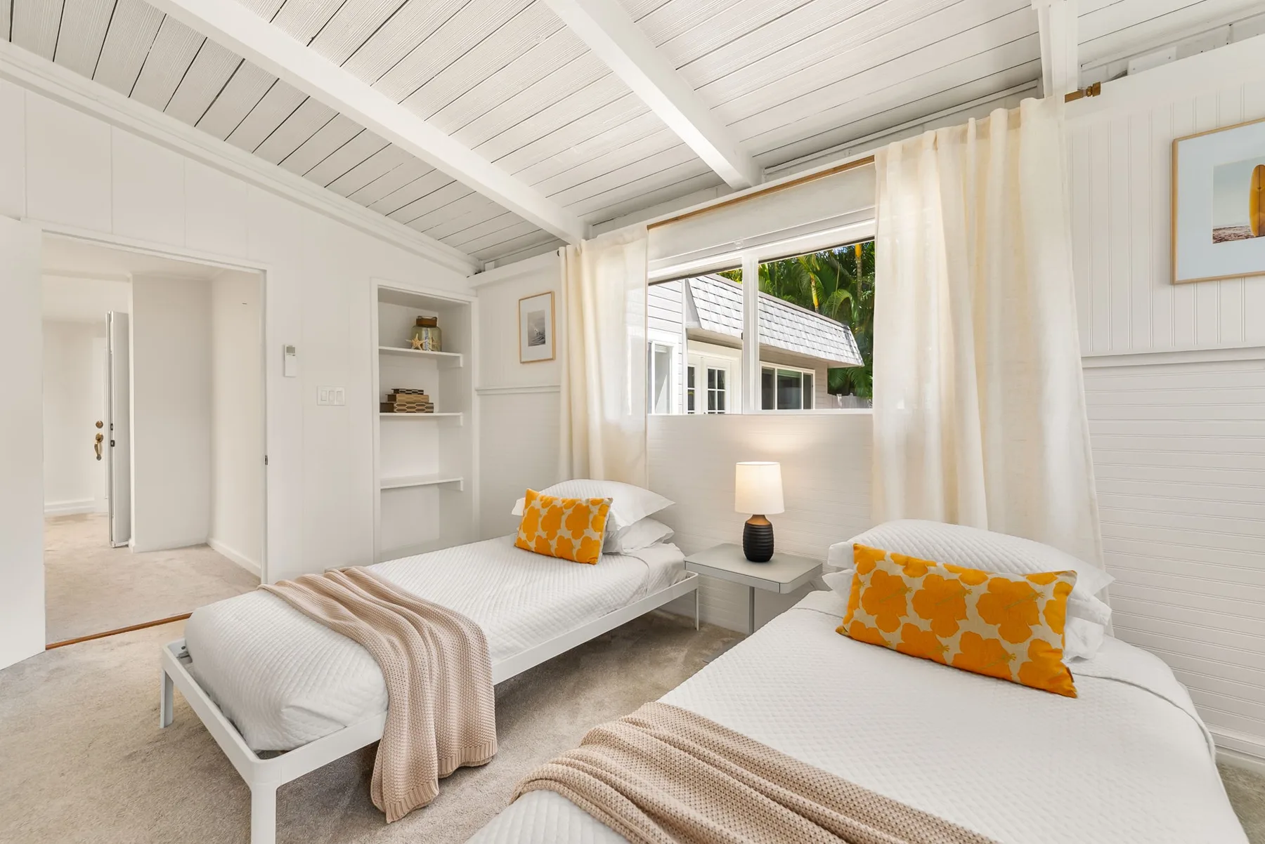 Un dormitorio luminoso con dos camas, cojines naranjas y cortinas blancas.