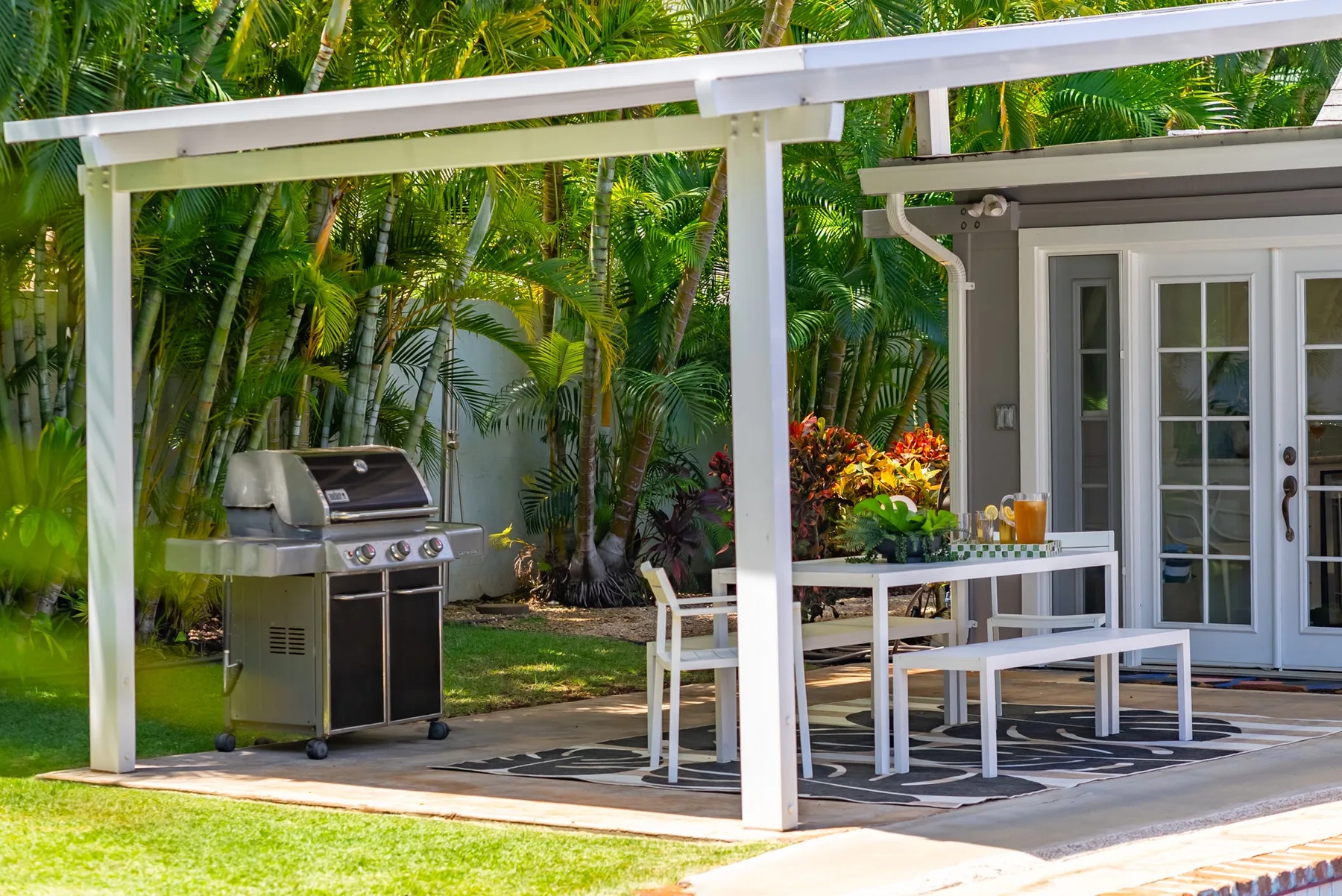 Un patio cubierto tiene una mesa de comedor junto a una parrilla y un jardín.
