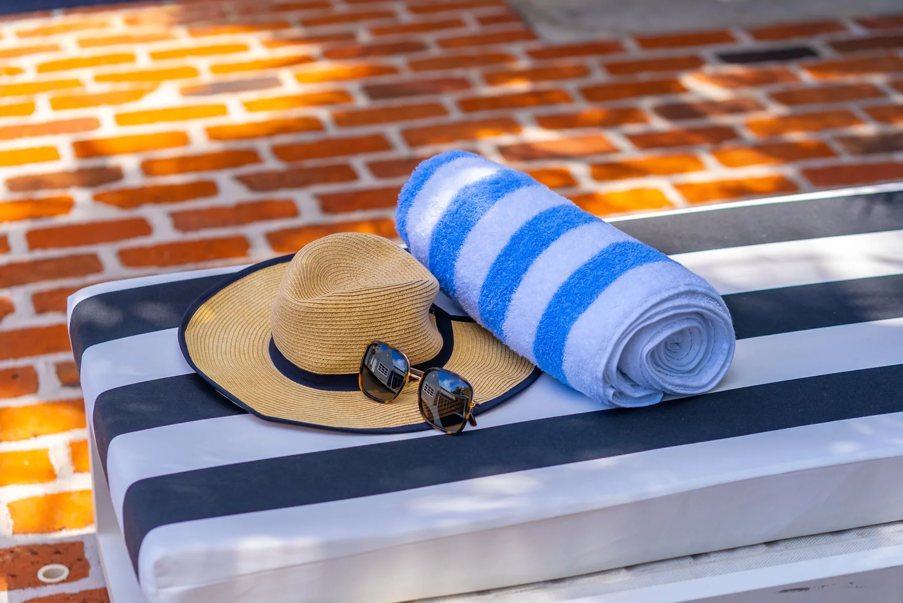 Un cojín de descanso a rayas sostiene un sombrero de paja, gafas de sol y una toalla azul enrollada.