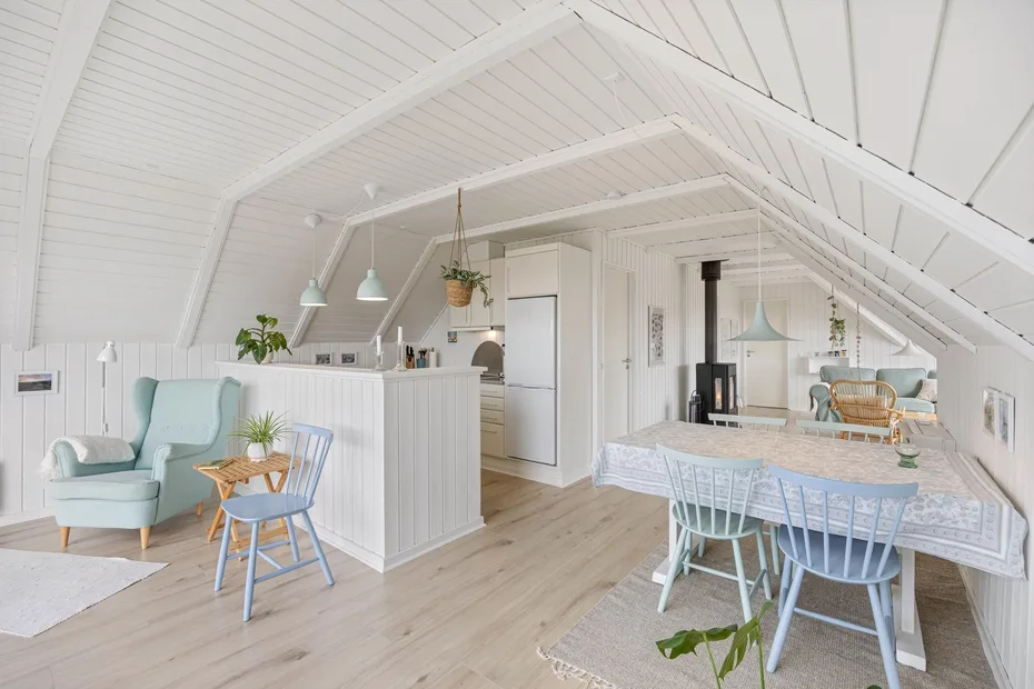 A bright open-plan dining and living area has white wood-paneled ceilings and pale furnishings.