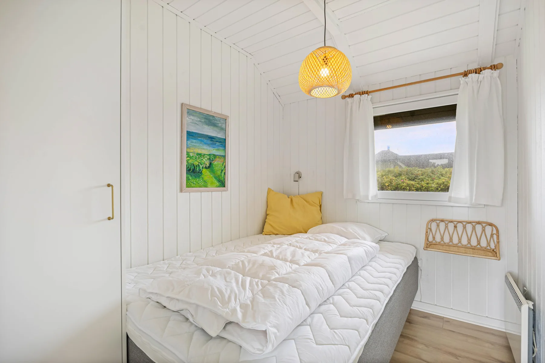 A bright bedroom with a bed, white bedding, and a window overlooking greenery.