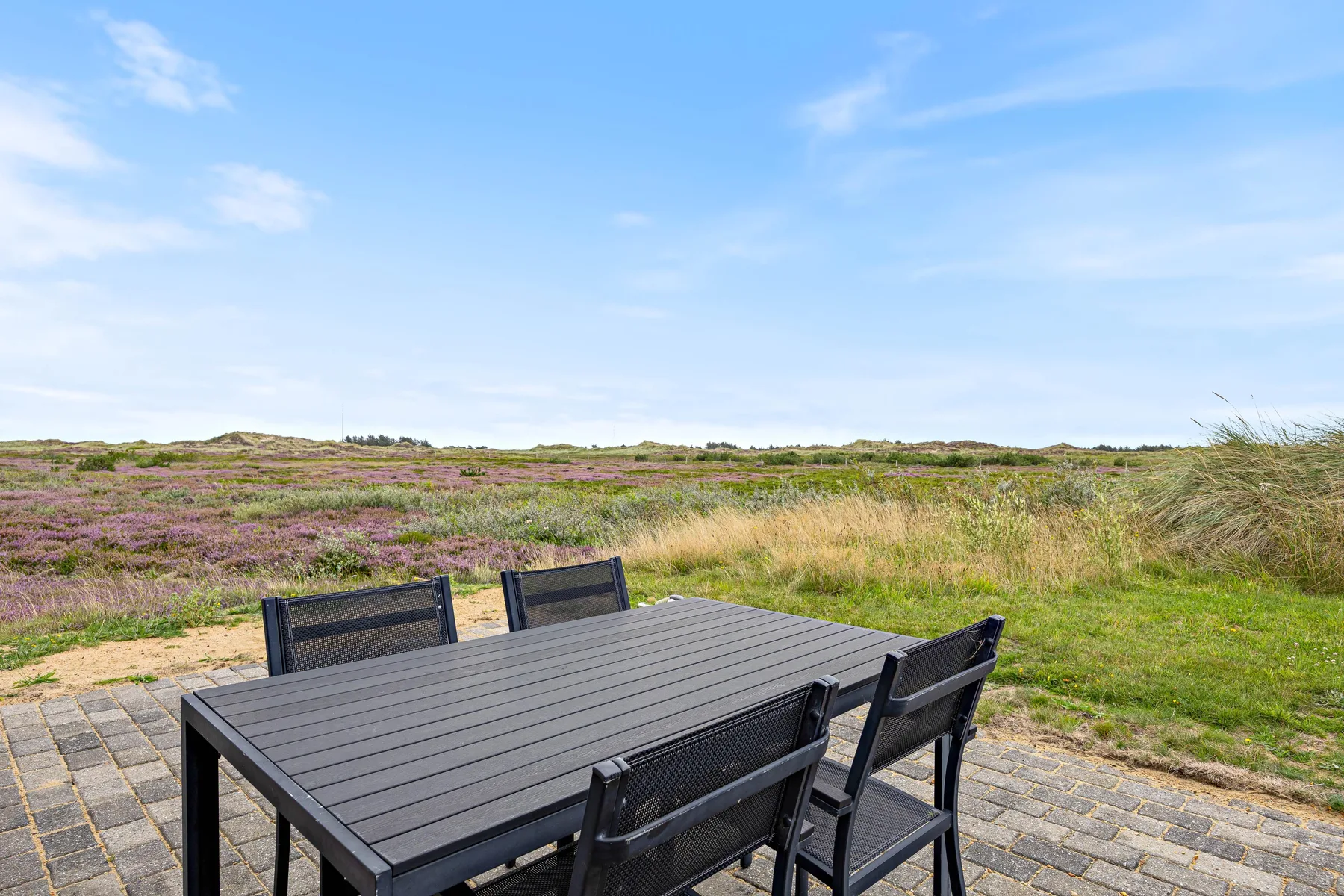 A patio dining table and seating overlook open heathland beneath a blue sky.