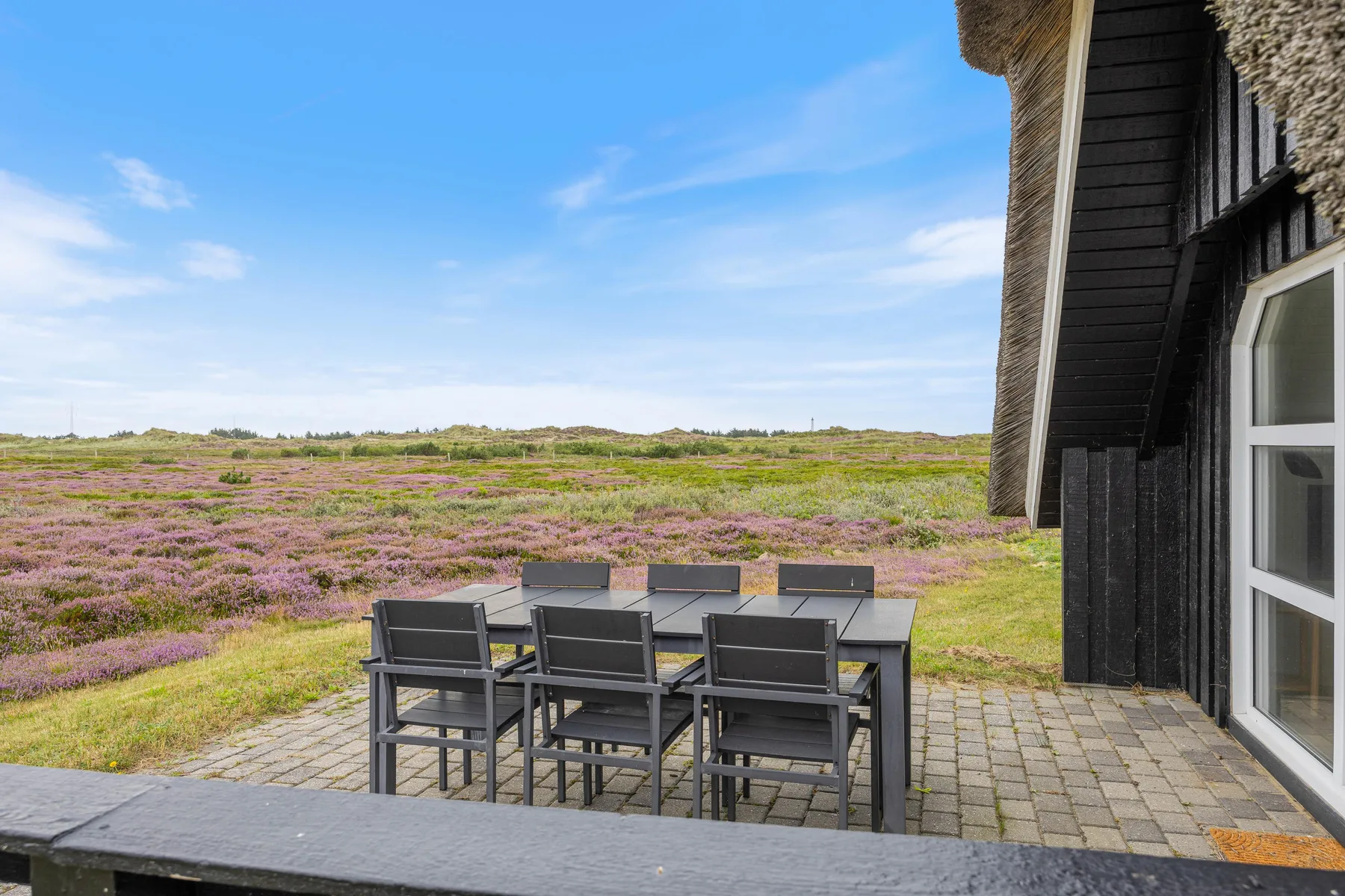 A patio seating area beside a dark house overlooks a field of purple flowers.