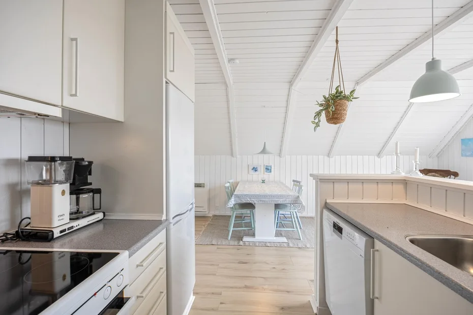A bright kitchen opens onto a dining area, with a long table beneath a hanging plant.