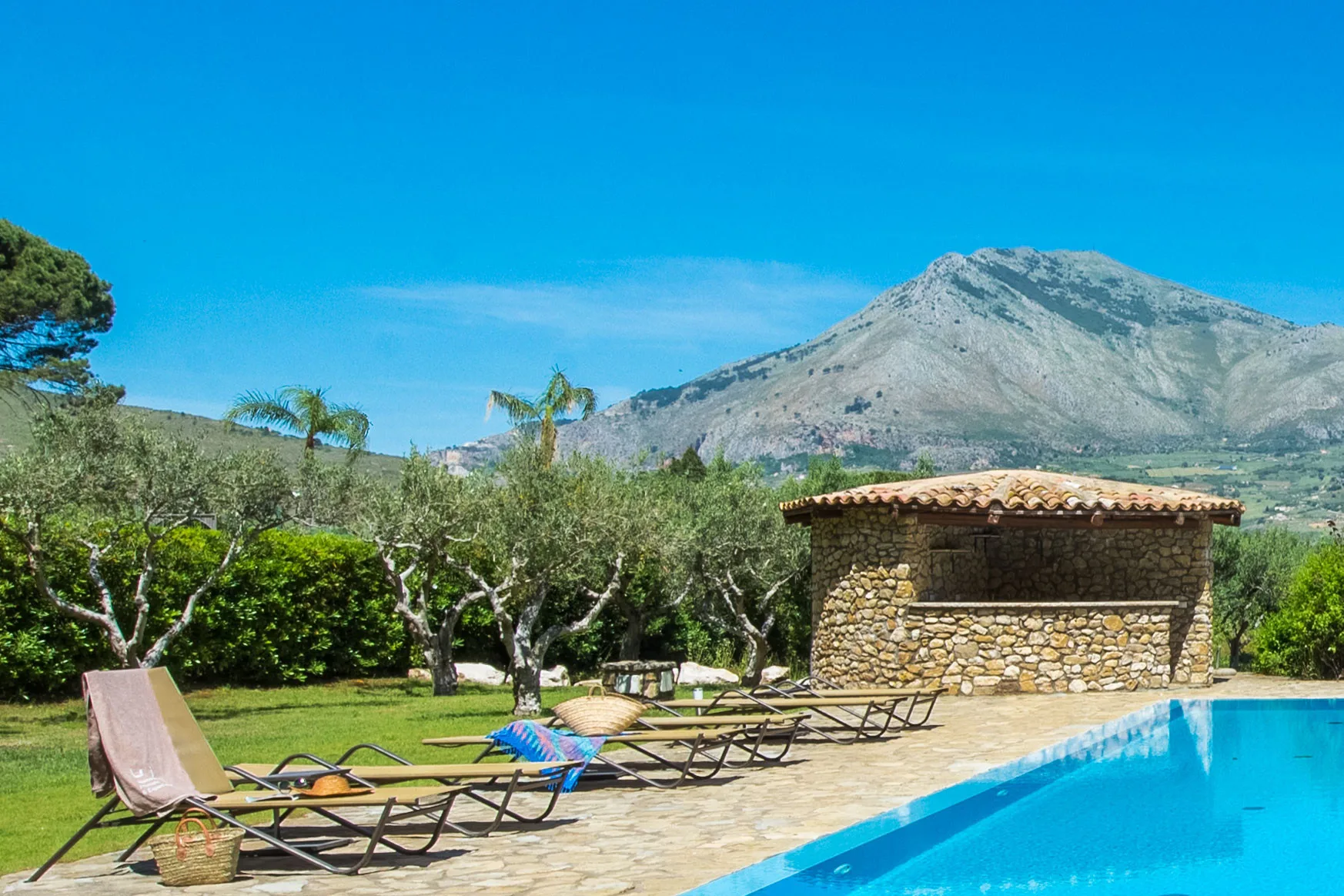 Una piscina y tumbonas están junto a un edificio de piedra, con una montaña al fondo.