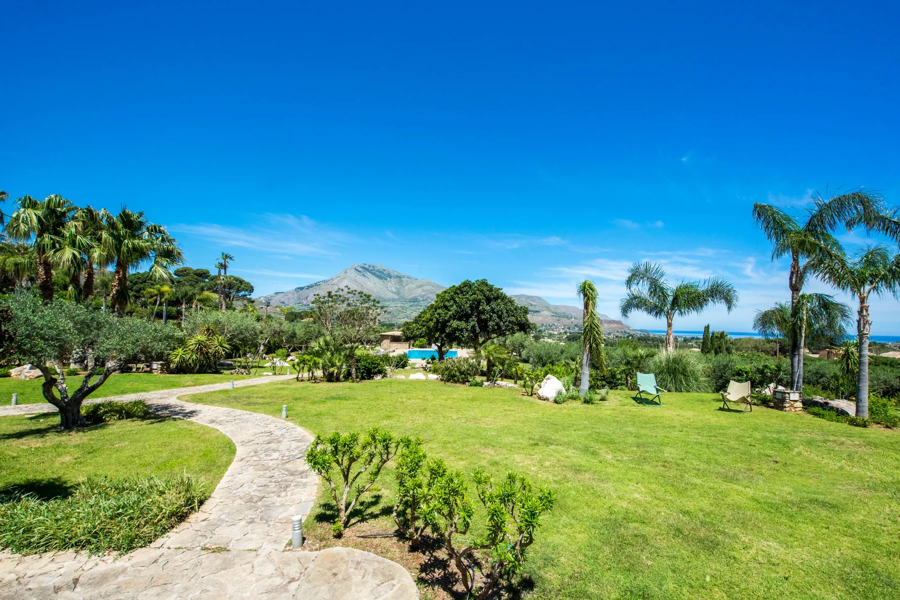 A landscaped garden with a curving path, palm trees, and distant hills.