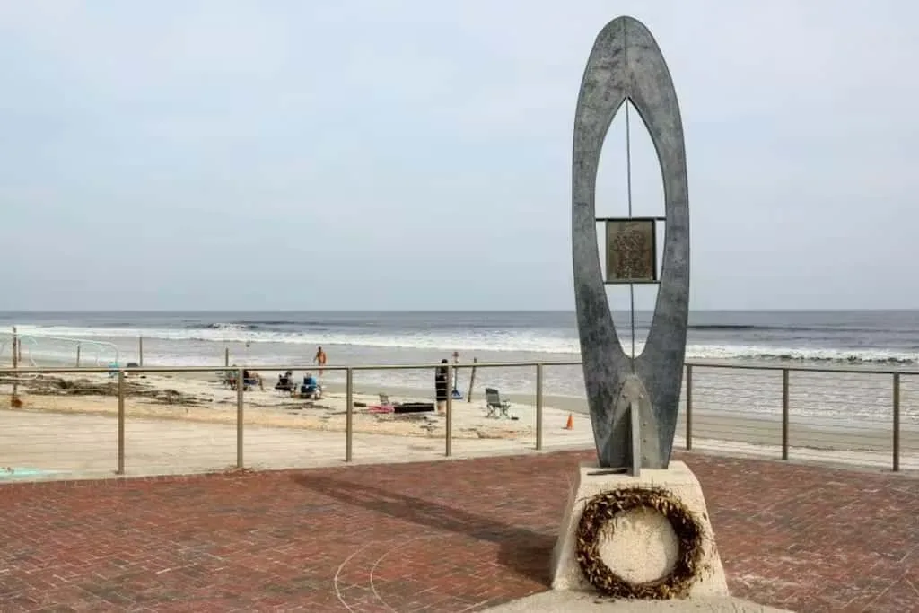 A seaside promenade overlooks the beach, with a tall sculptural monument in the foreground.