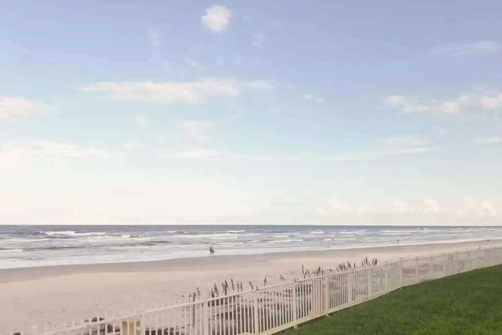 A sandy beach meets the ocean beside a grassy dune and fence.