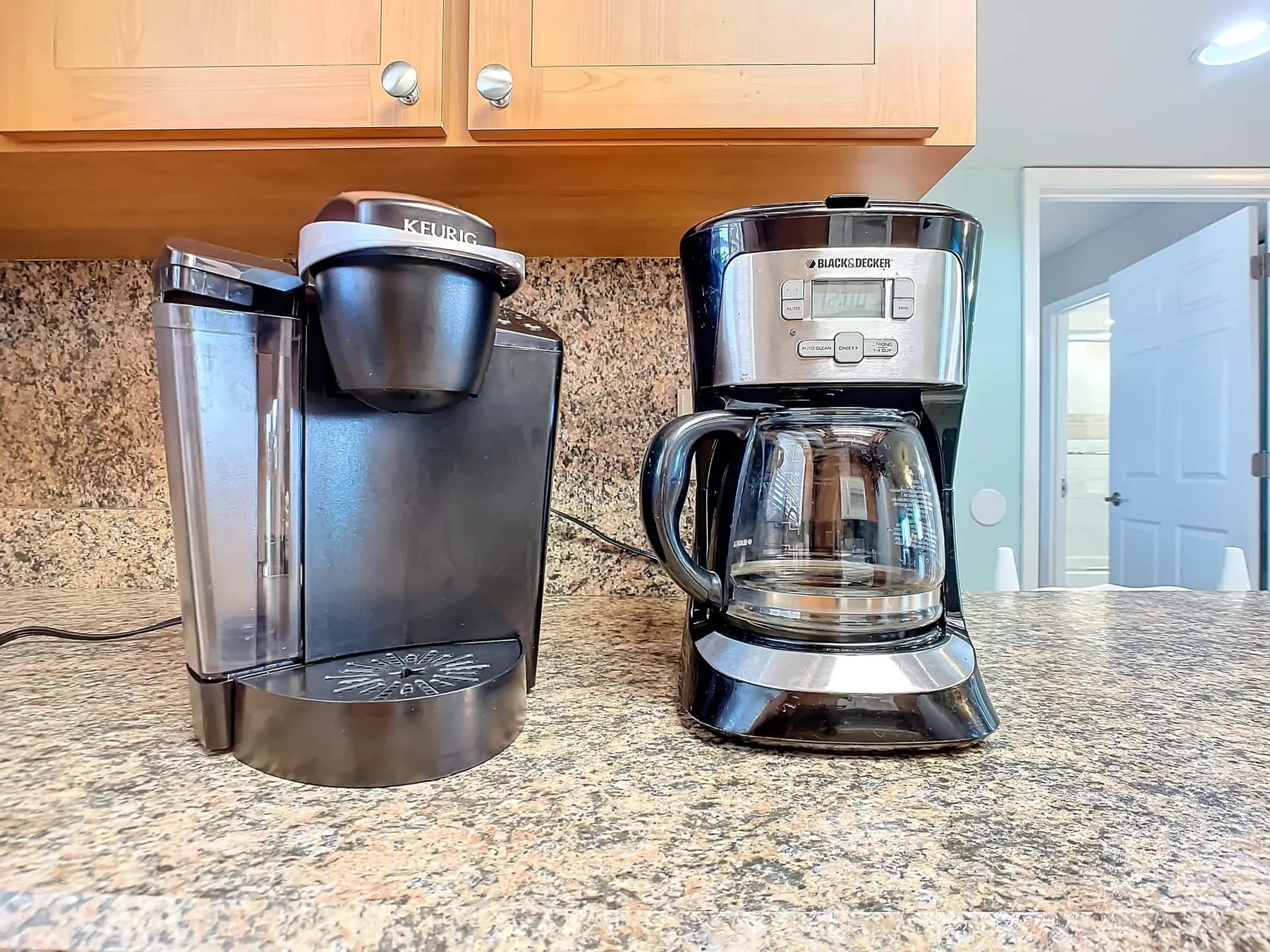 A kitchen counter holds two coffee makers beneath wooden cabinets.