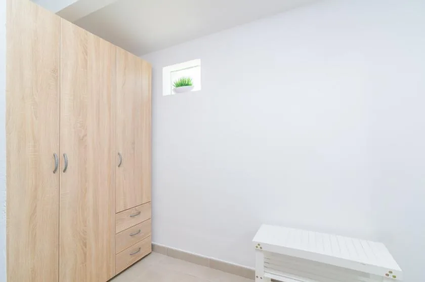 Una habitación luminosa con un armario de madera, un banco blanco y una pequeña ventana alta.