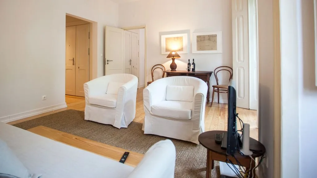 Una sala luminosa con dos sillones blancos, una alfombra y un televisor pequeño.
