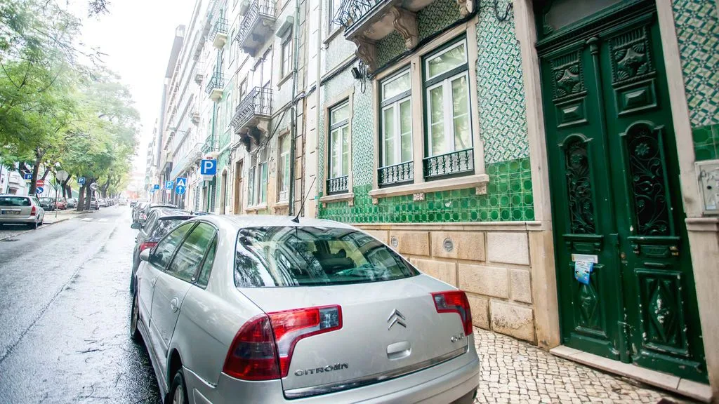 Una calle urbana con edificios, coches estacionados y una entrada verde.