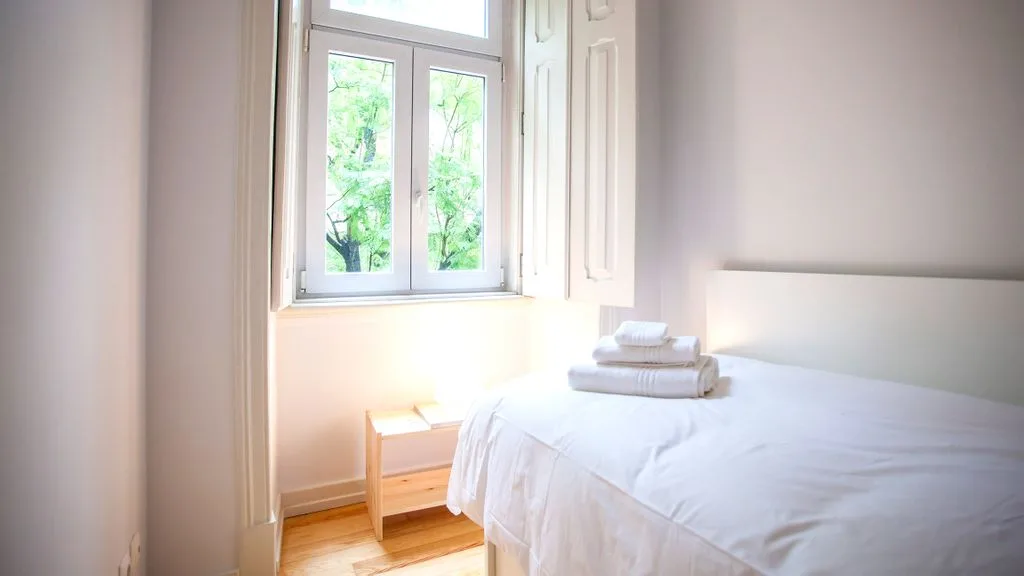 Un dormitorio luminoso con ropa de cama blanca junto a una ventana alta.