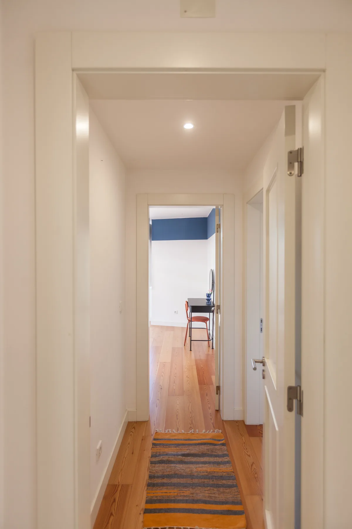 A narrow hallway with wooden flooring and a striped runner leads to another room.