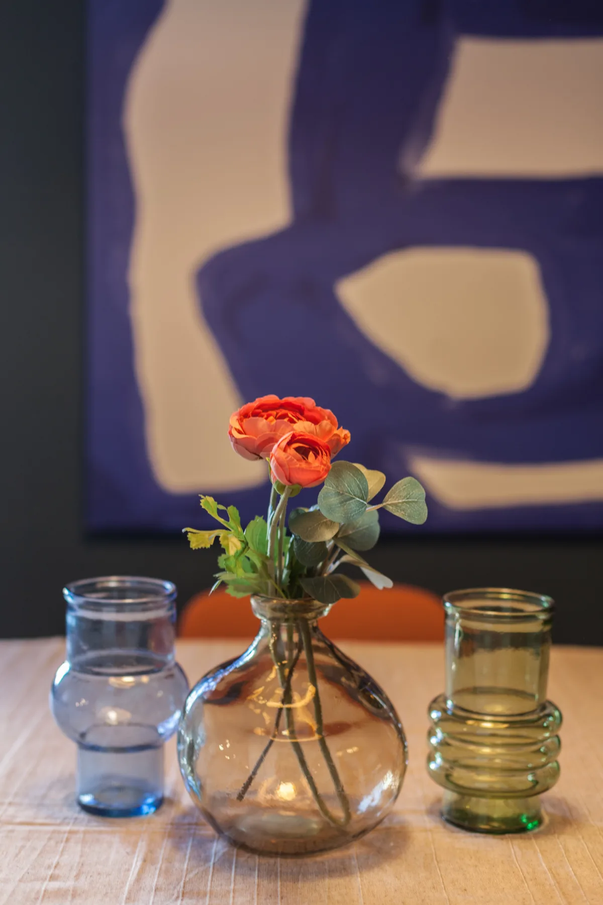 A table with a glass vase of flowers and drinking glasses in front of abstract artwork.