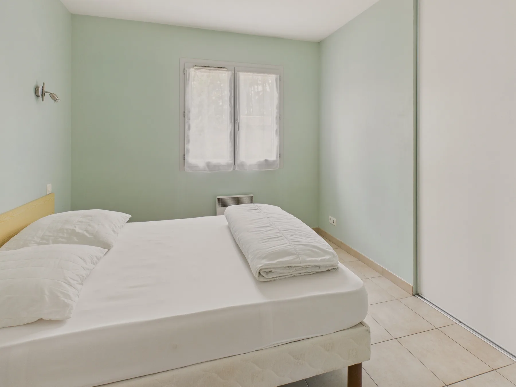 A bedroom with a bed, pale green walls, and a small curtained window.