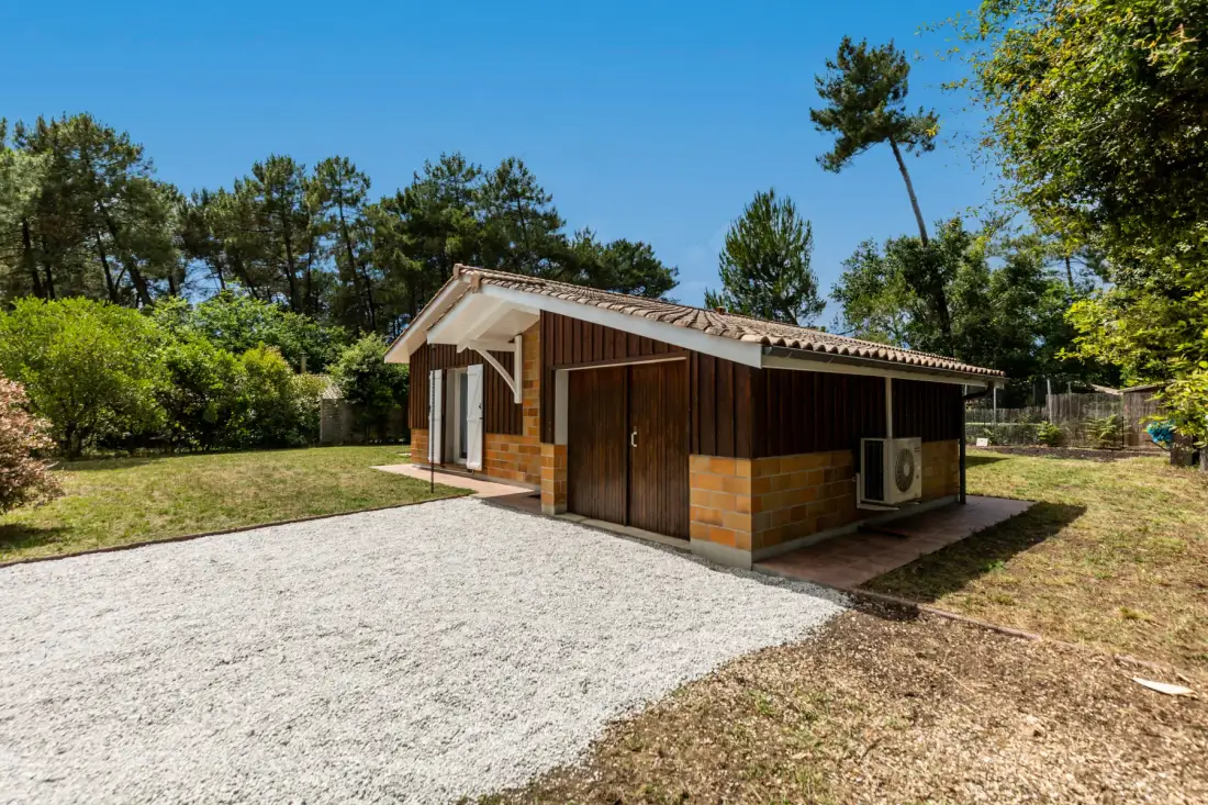 A small house stands beside a gravel driveway in a tree-lined yard.