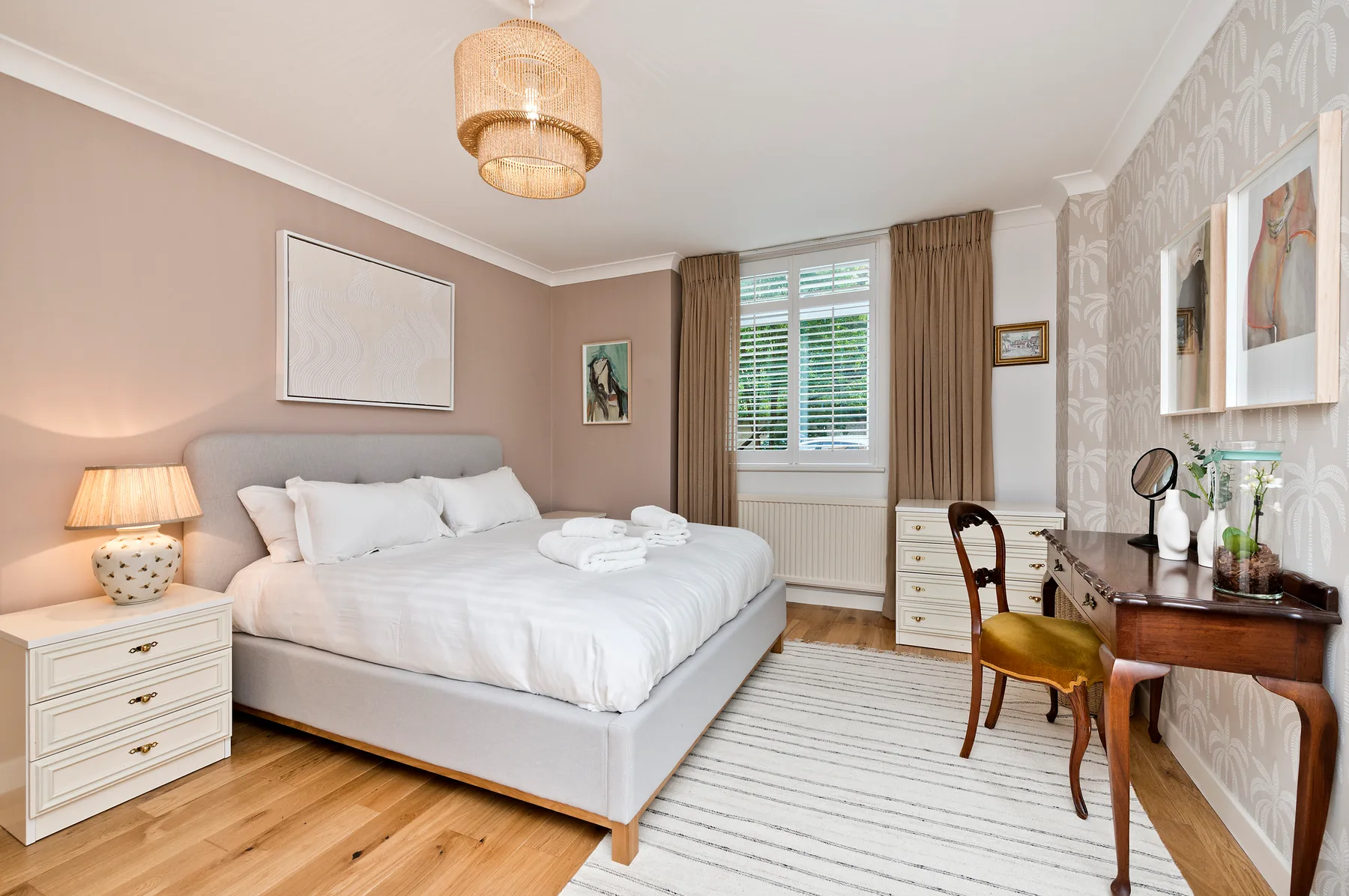 A bright bedroom with a neatly made bed, a desk, and a window with blinds.