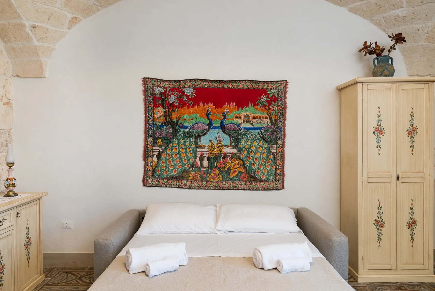 A bedroom with a bed beneath a colorful woven wall hanging.
