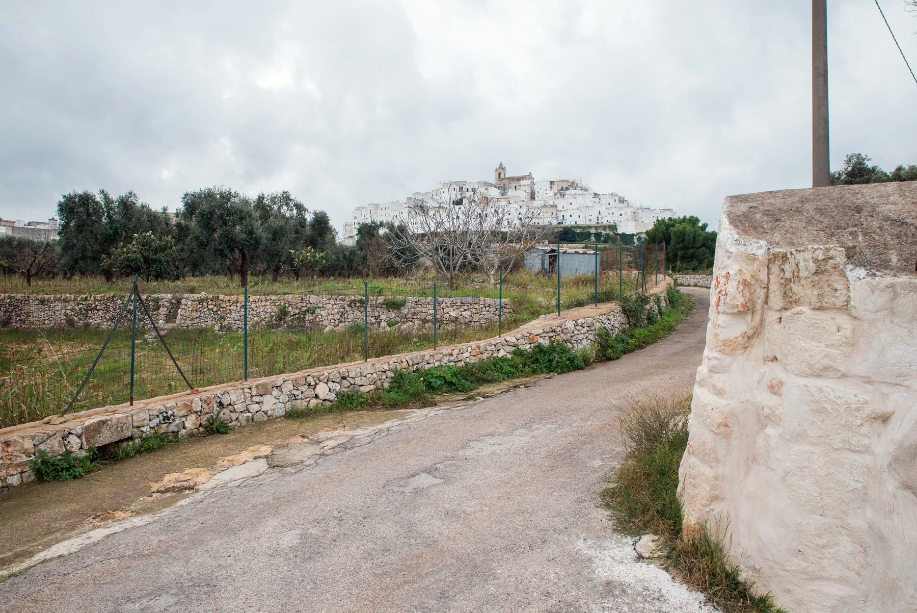 A country road curves past a stone wall, with a hilltop village in the distance.