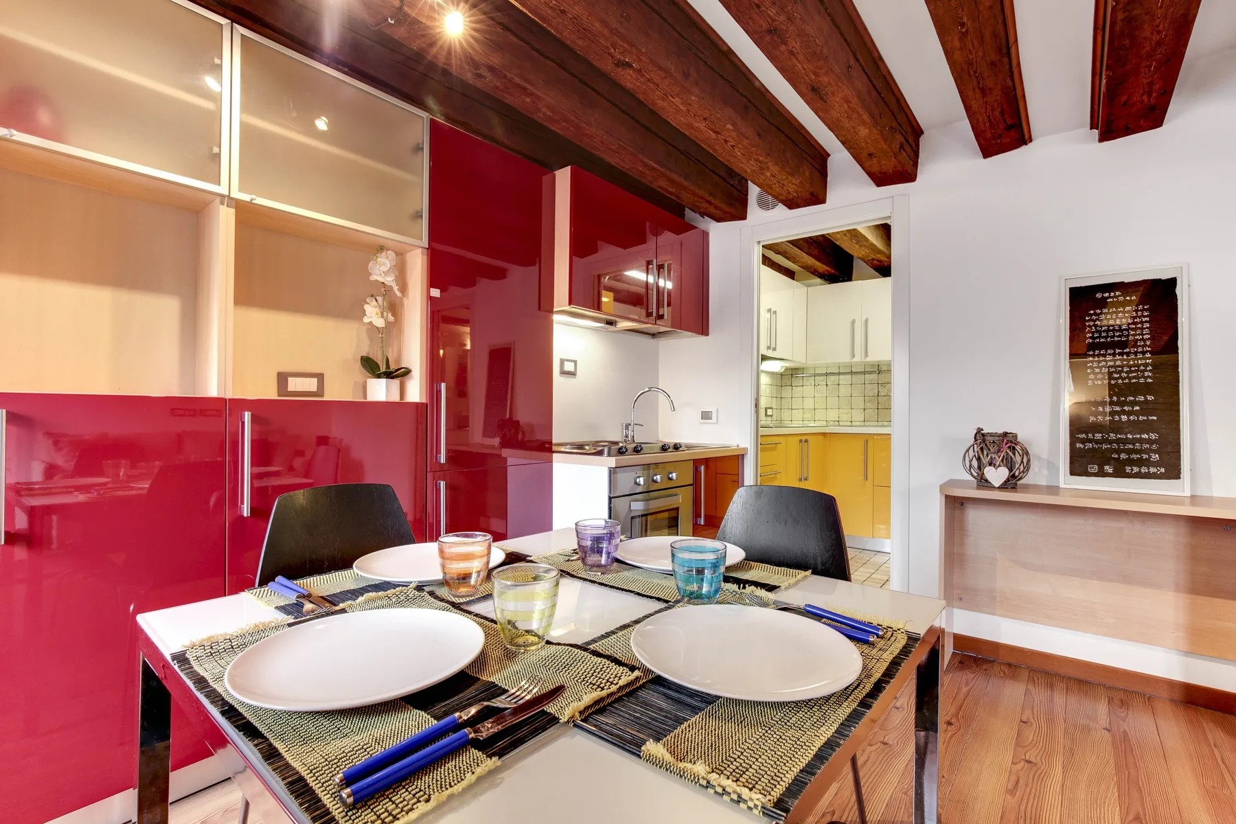 A dining table is set with plates and glasses beside red kitchen cabinets.