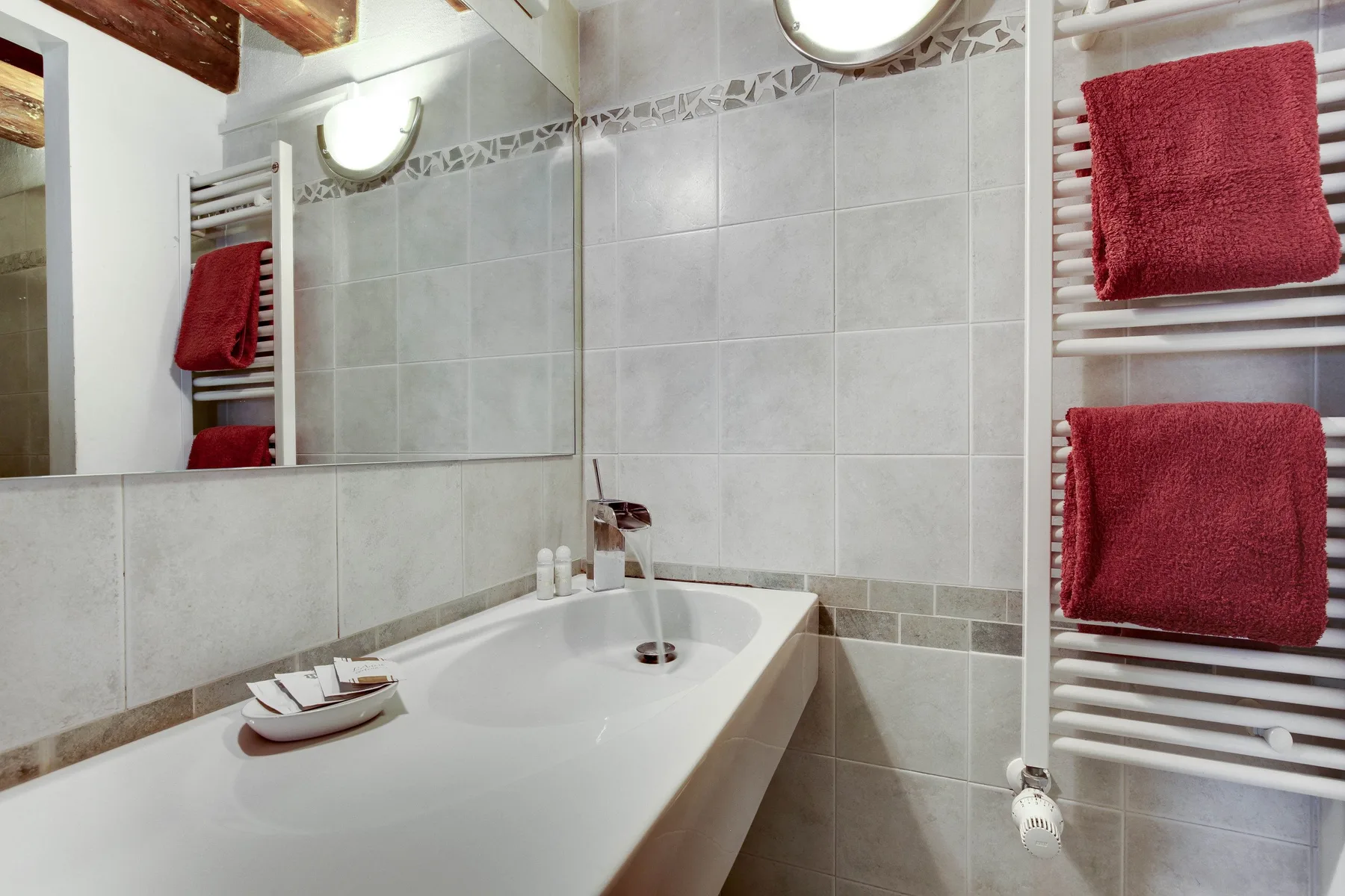 A tiled bathroom with a sink, wall mirror, and red towels on a heated towel rail.