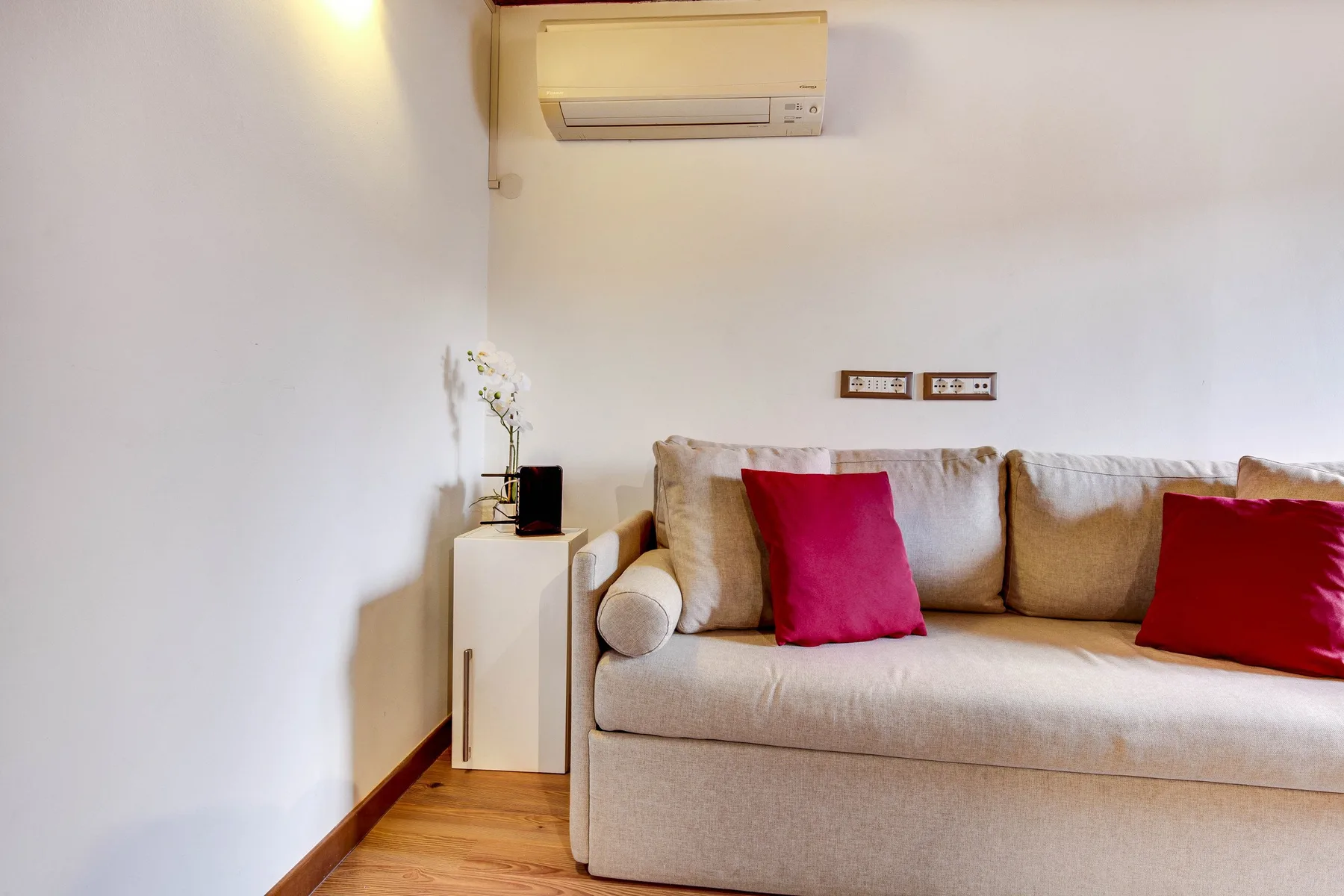 A living room with beige seating and red cushions beneath a wall-mounted air conditioner.
