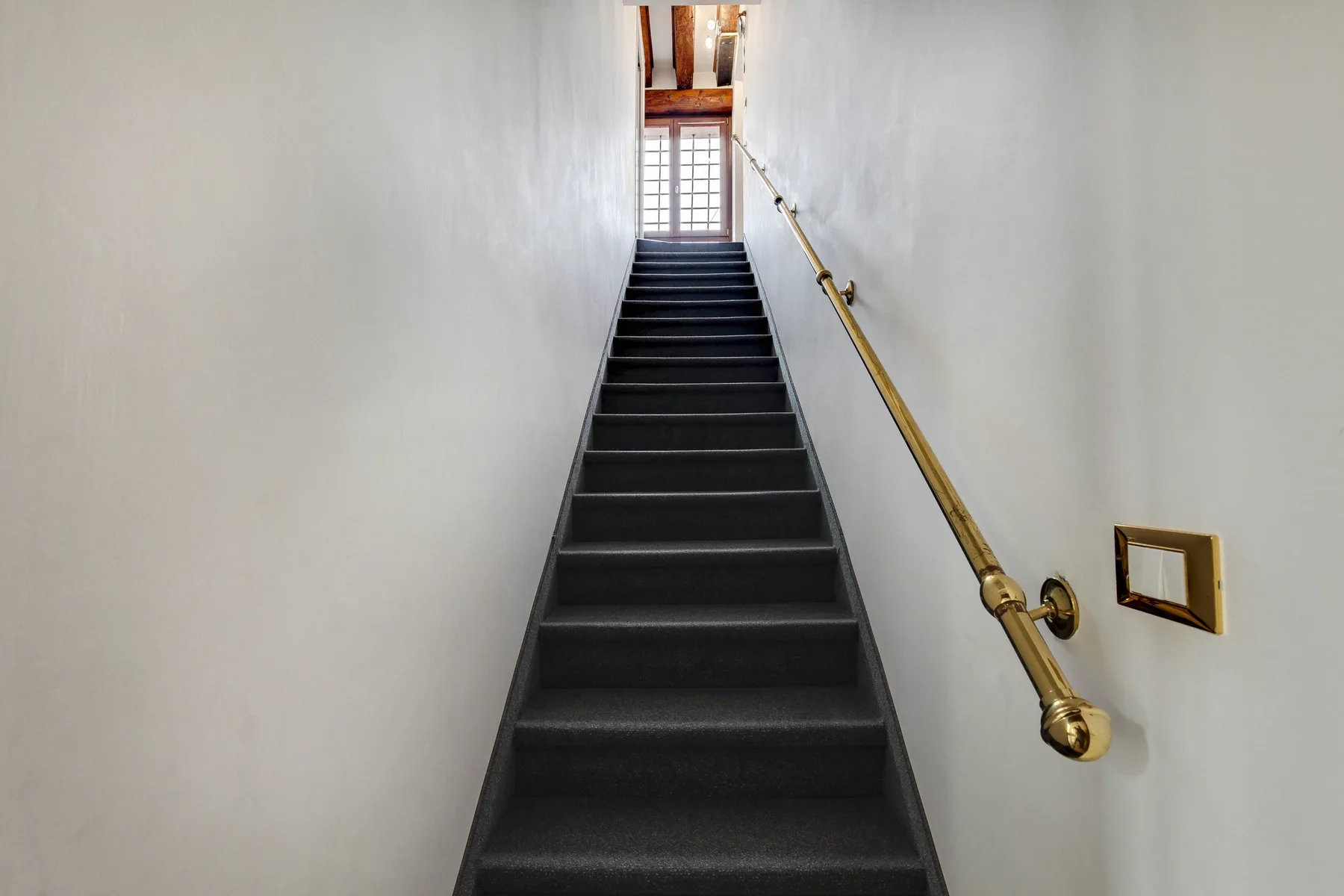A narrow staircase with dark steps and a brass handrail leads to a window.