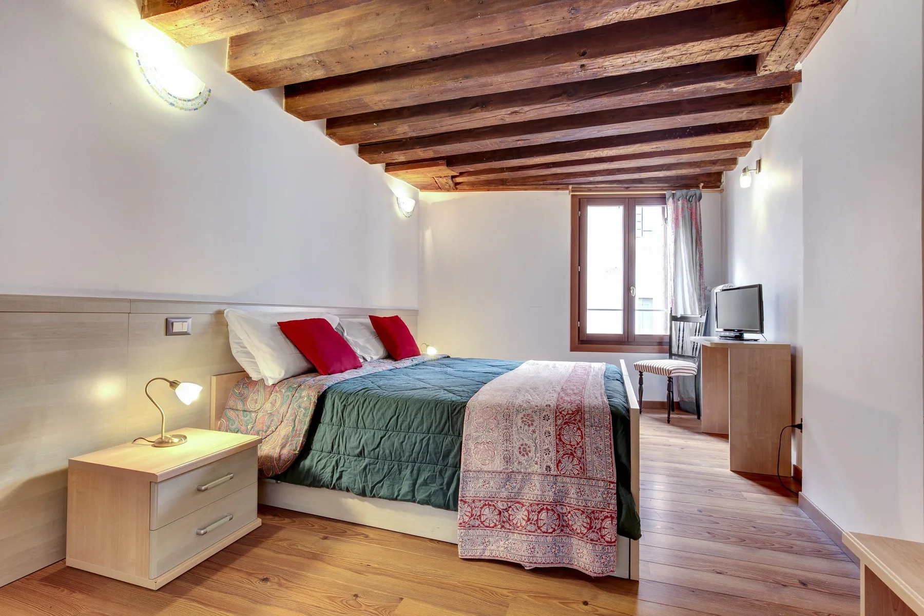 A bedroom with a double bed, wooden ceiling beams, and a window.