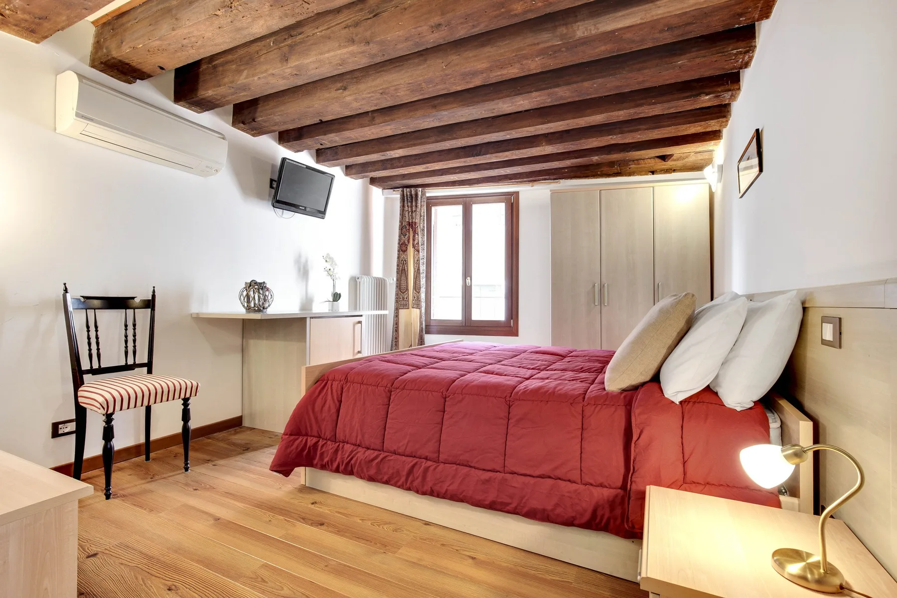 A bedroom with a red bedspread, wooden ceiling beams, and a window.