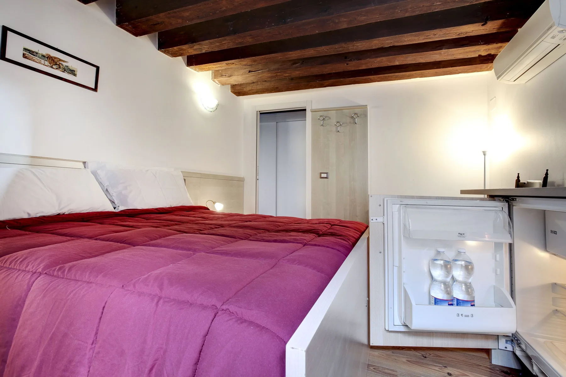 A bedroom with a bed covered by a magenta quilt and dark exposed ceiling beams.