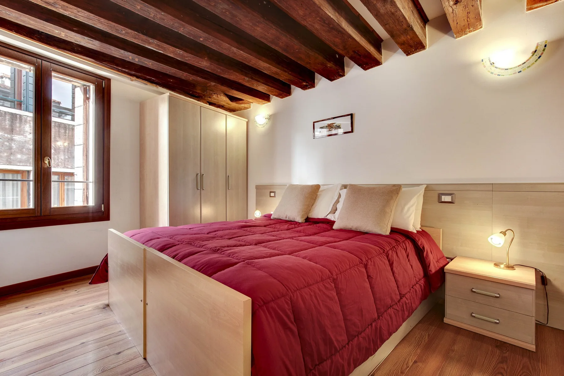 A bedroom with a bed covered in red bedding beside a large window.