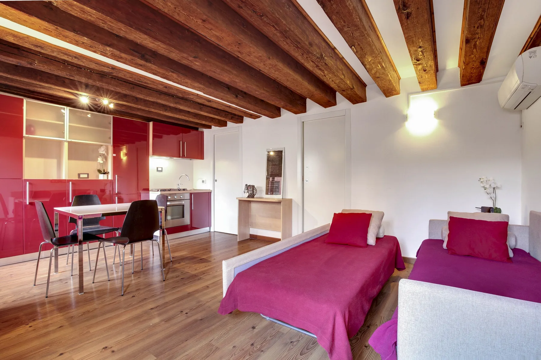 A bedroom with two beds and a compact kitchen area beneath exposed wooden beams.