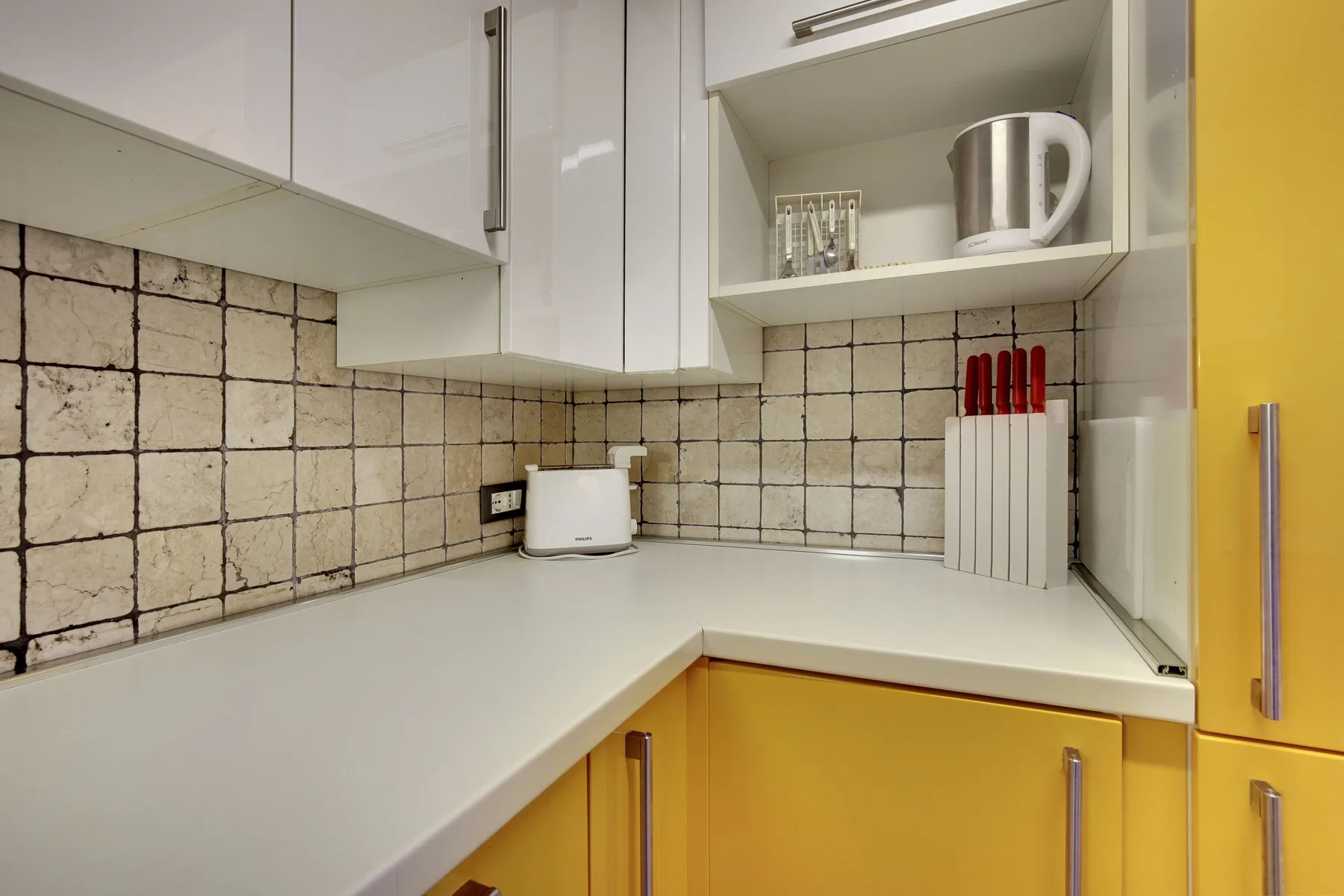 A kitchen corner with white countertops, yellow cabinets, and tiled walls.
