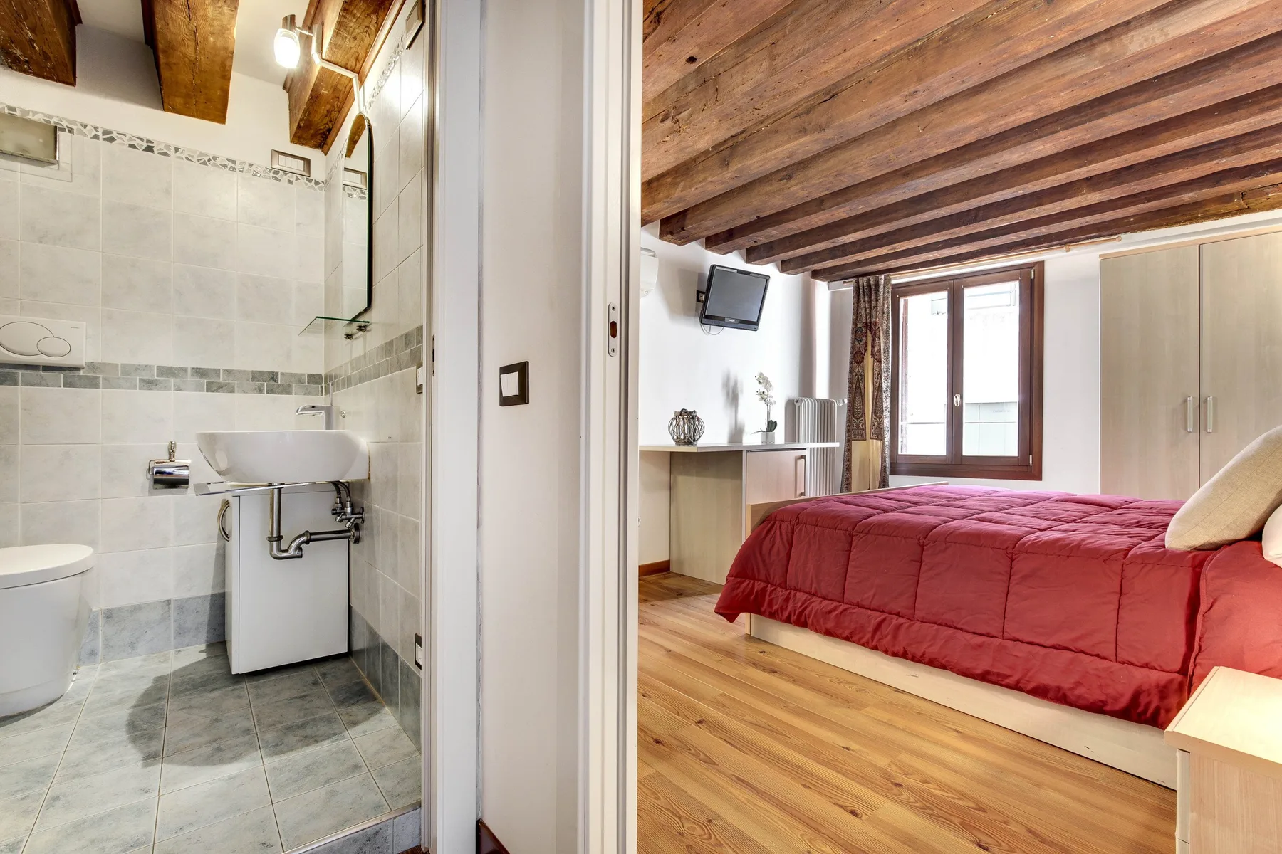 A bedroom with a red bedspread sits beside a tiled bathroom.