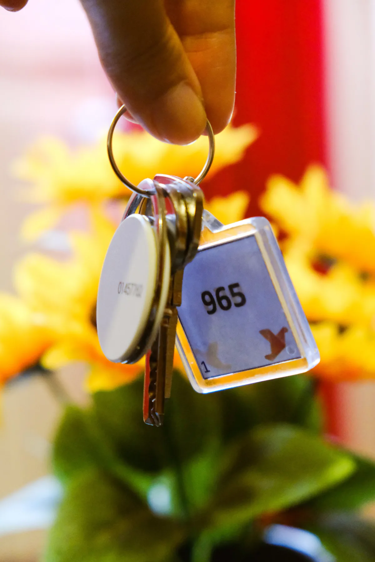 A hand holds a set of keys with a numbered tag against a blurred floral background.