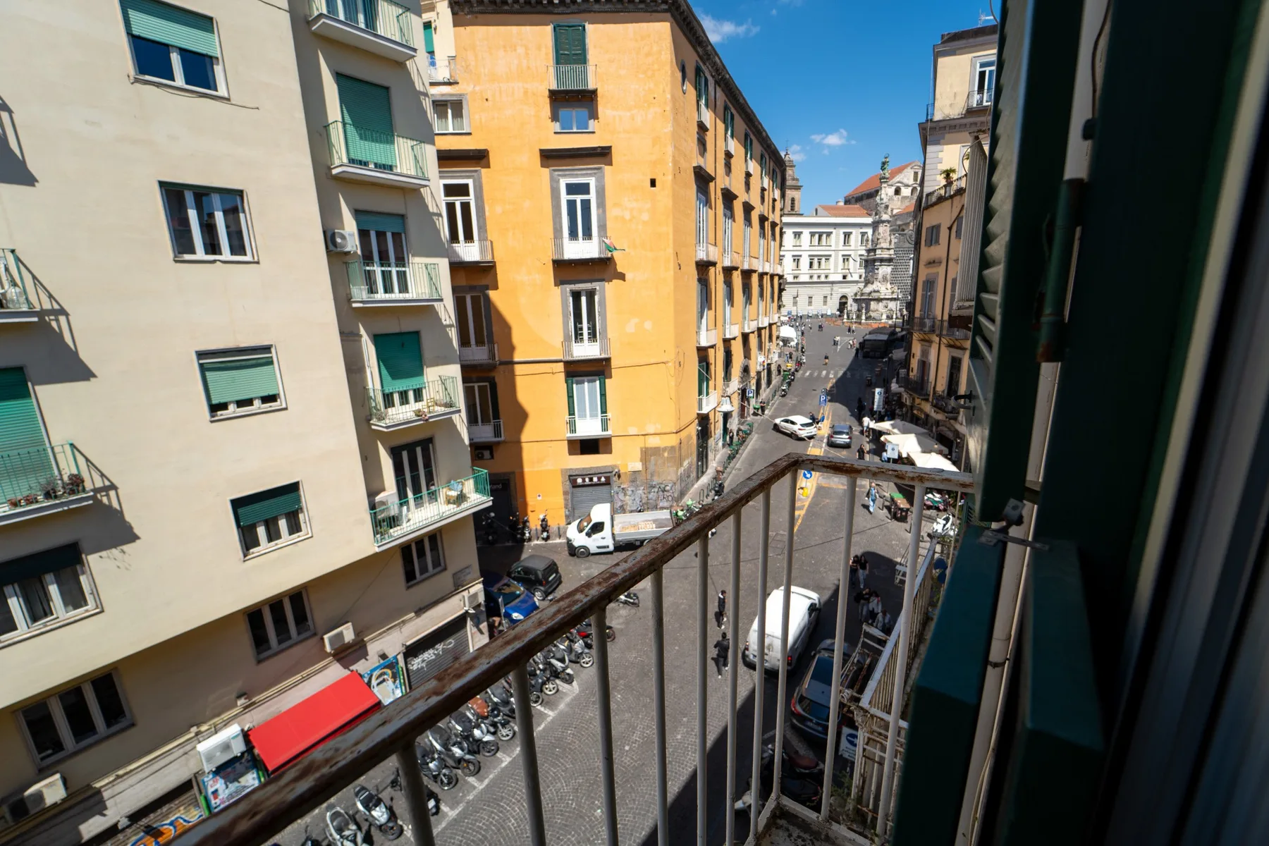 A city street viewed from a balcony, with apartment buildings and parked vehicles.