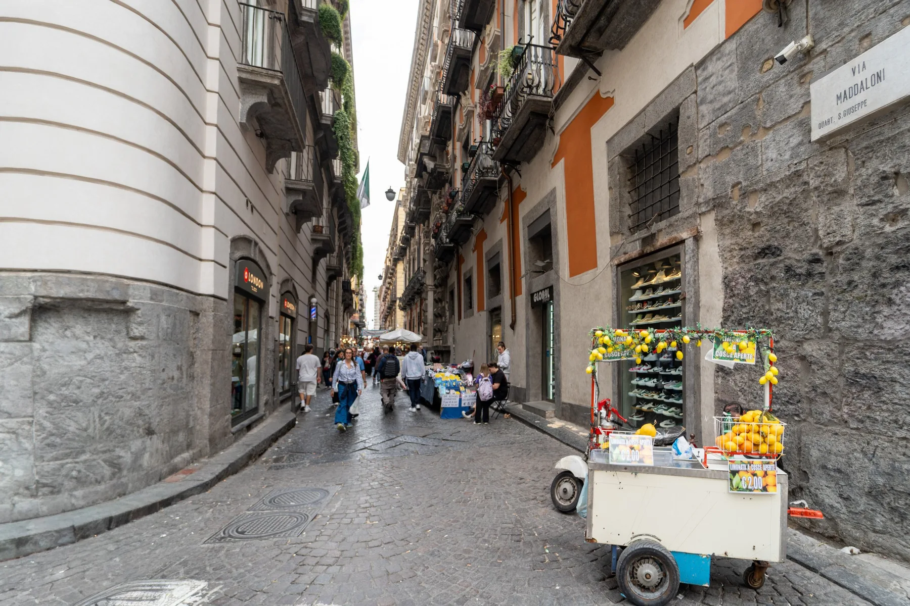 A narrow cobblestone street lined with buildings, with a small produce cart in the foreground.