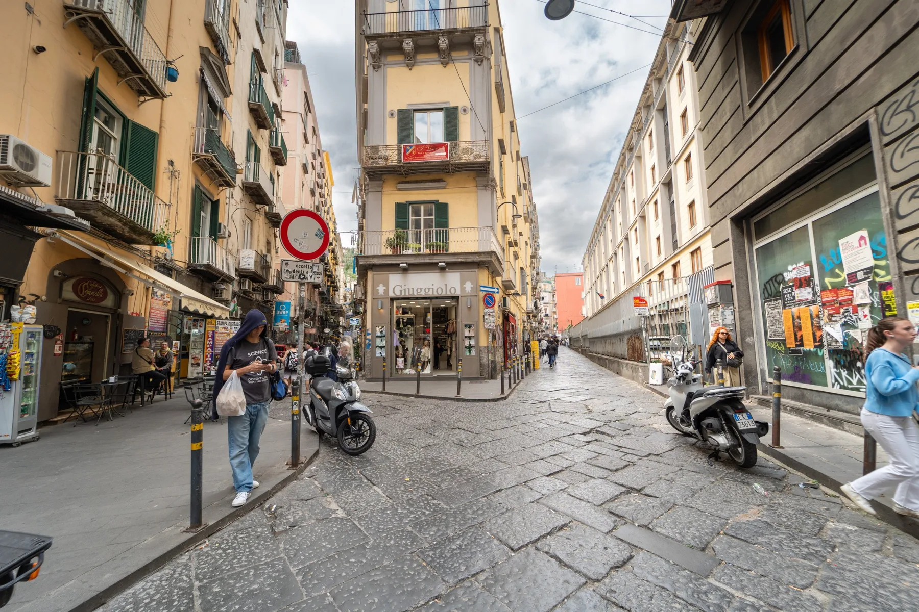 A narrow city street lined with tall buildings, shops, and parked scooters.