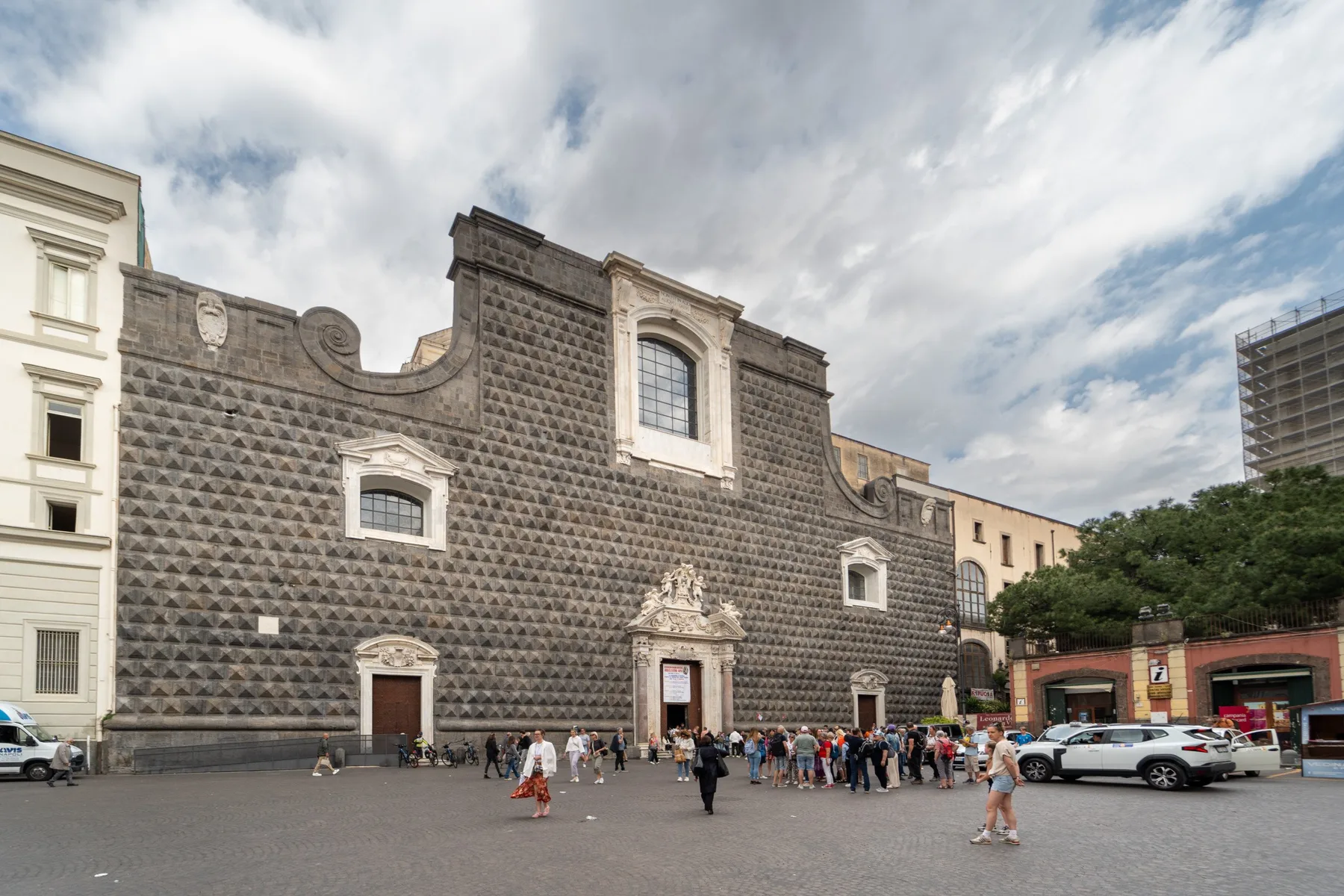 A large historic stone building faces an open plaza under a cloudy sky.