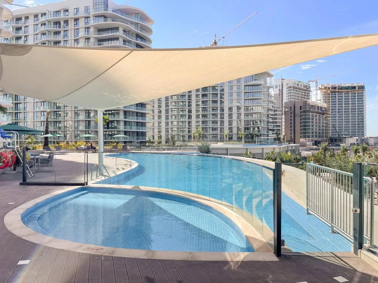 Una piscina exterior curva está bajo una gran cubierta de sombra, con edificios altos al fondo.