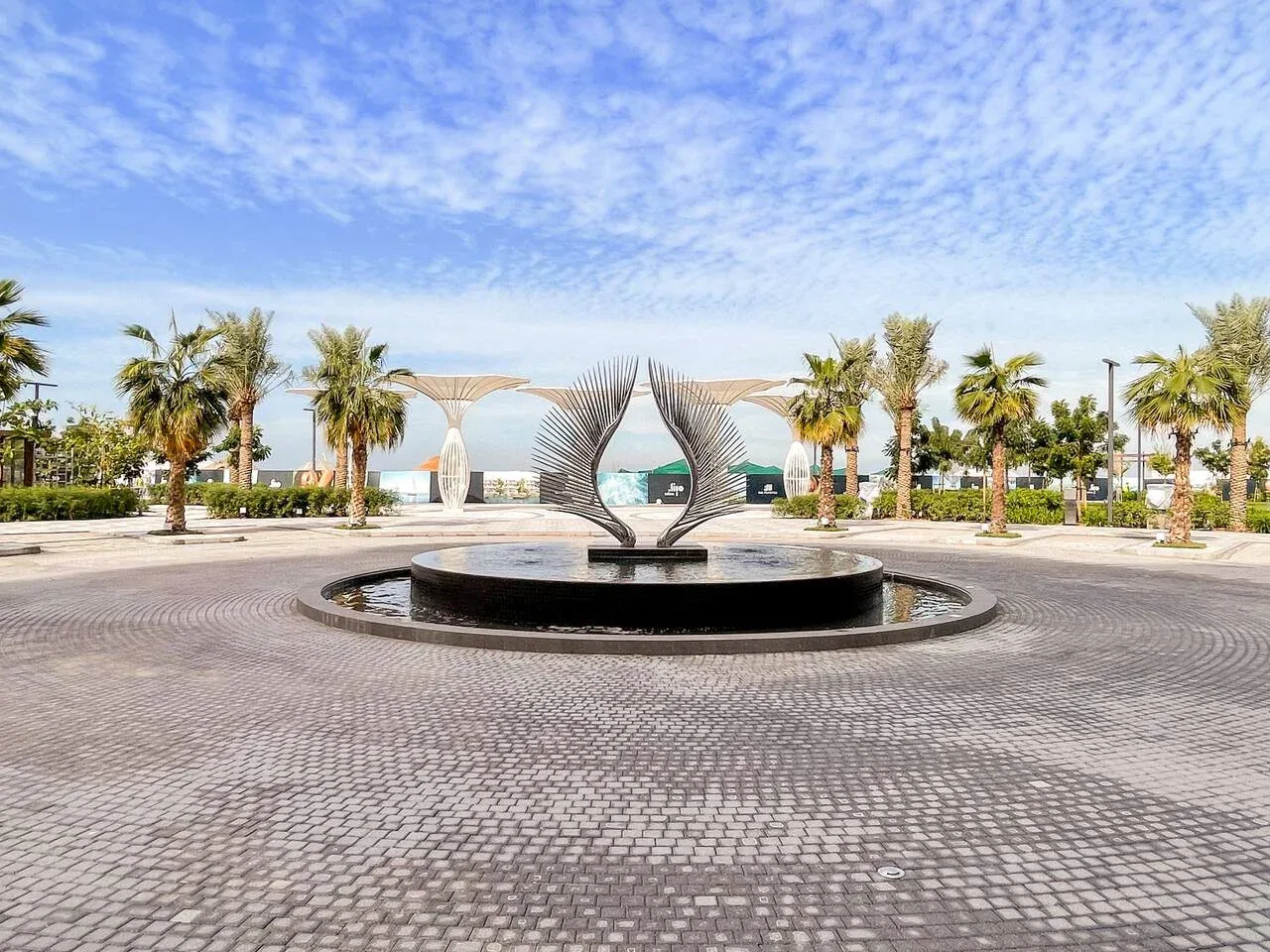 Una plaza pavimentada al aire libre tiene una fuente escultórica, con palmeras y el mar al fondo.