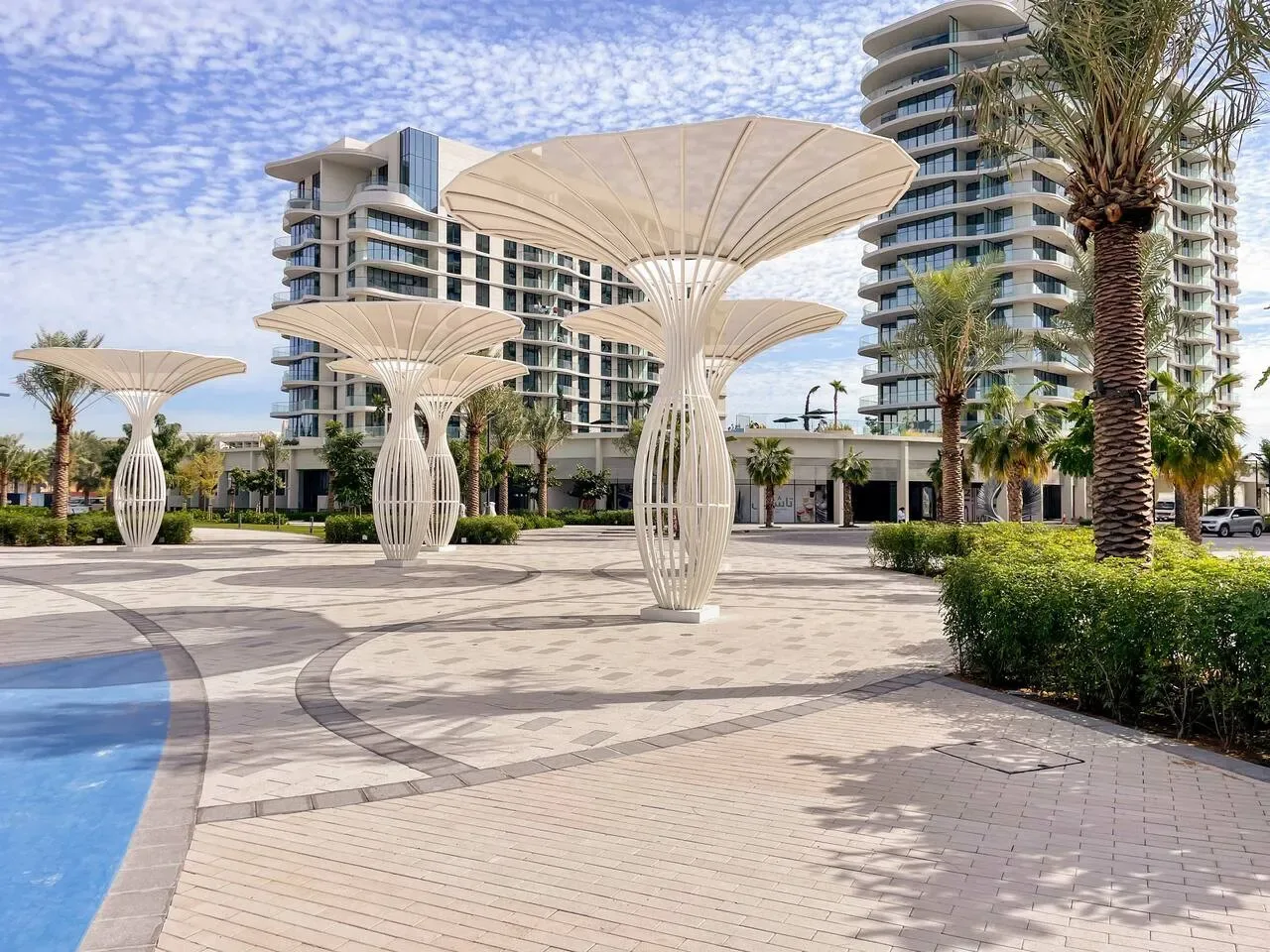 Una plaza exterior pavimentada tiene estructuras escultóricas de sombra junto a una piscina y edificios modernos.
