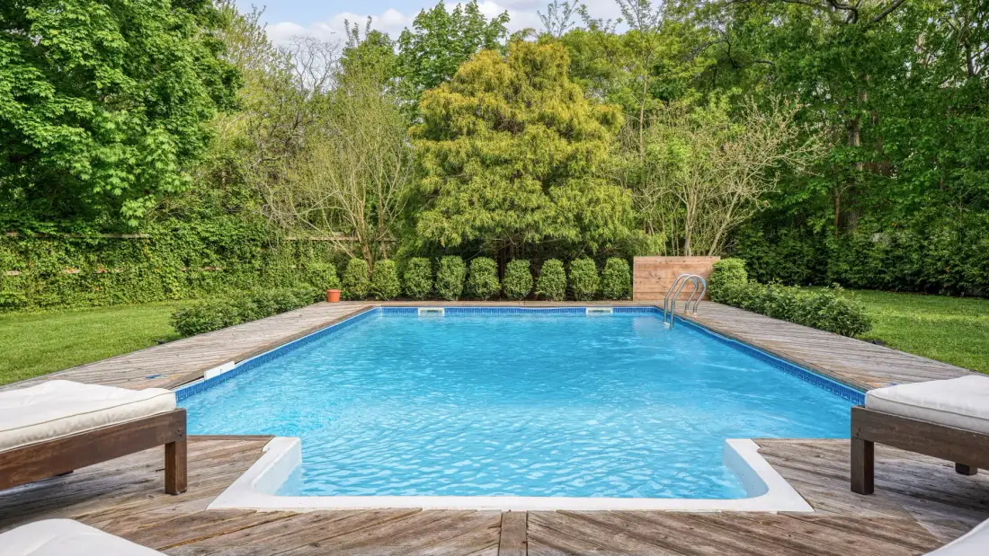 Una piscina al aire libre está rodeada por una terraza de madera y árboles frondosos.