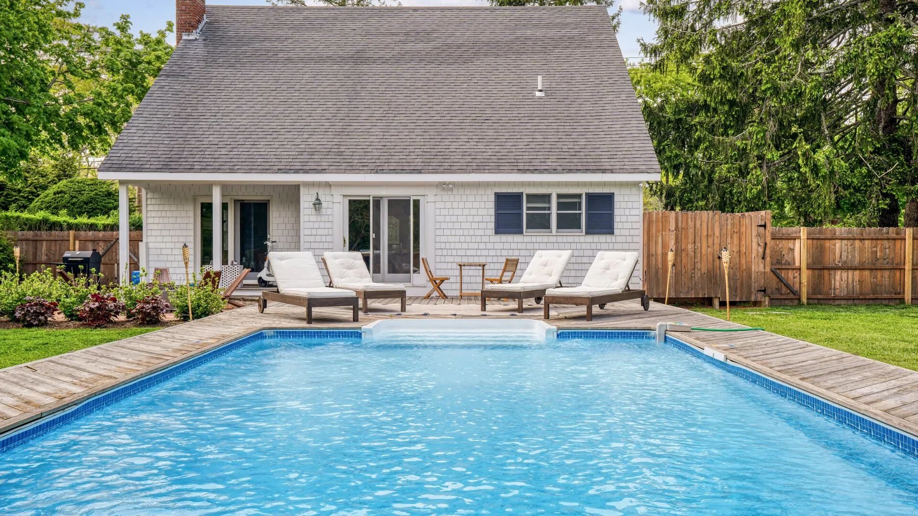 A backyard swimming pool sits beside a house and wooden deck.