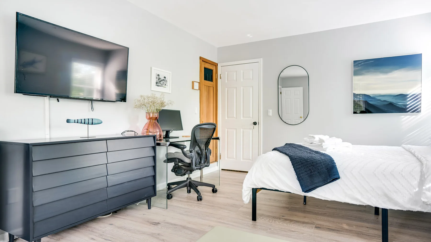 Un dormitorio con una cama, una cómoda y un escritorio bajo un televisor en la pared.