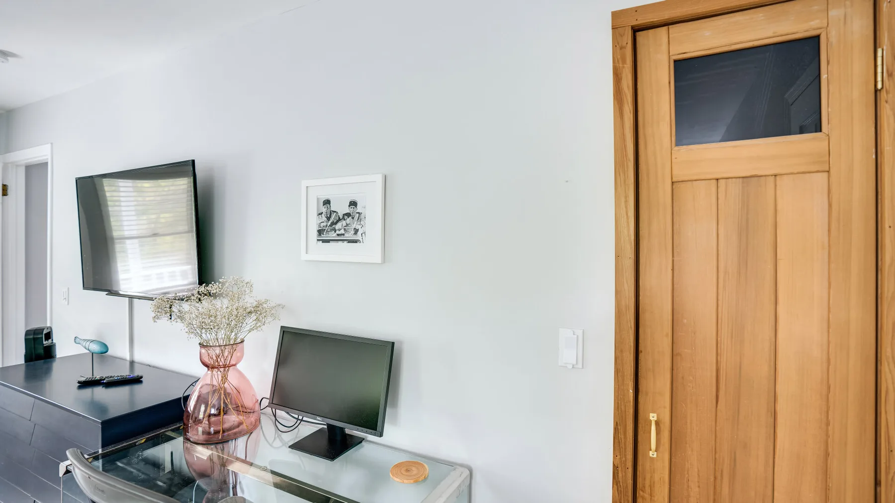 Un espacio interior luminoso tiene un escritorio de vidrio, un monitor y una puerta de madera.