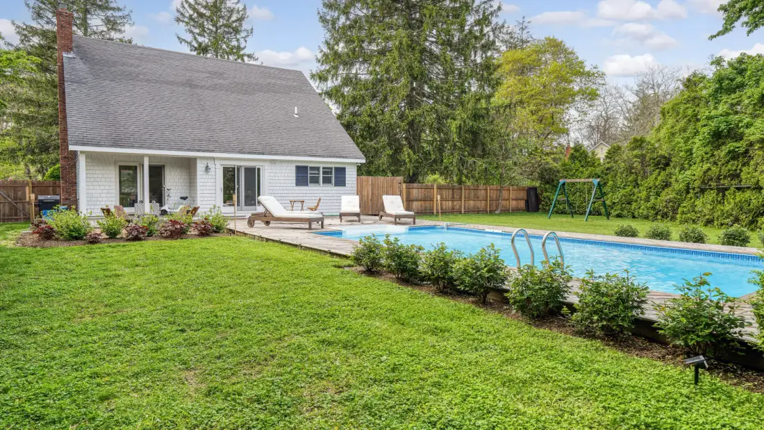 Una casa junto a una piscina, con césped y árboles alrededor.