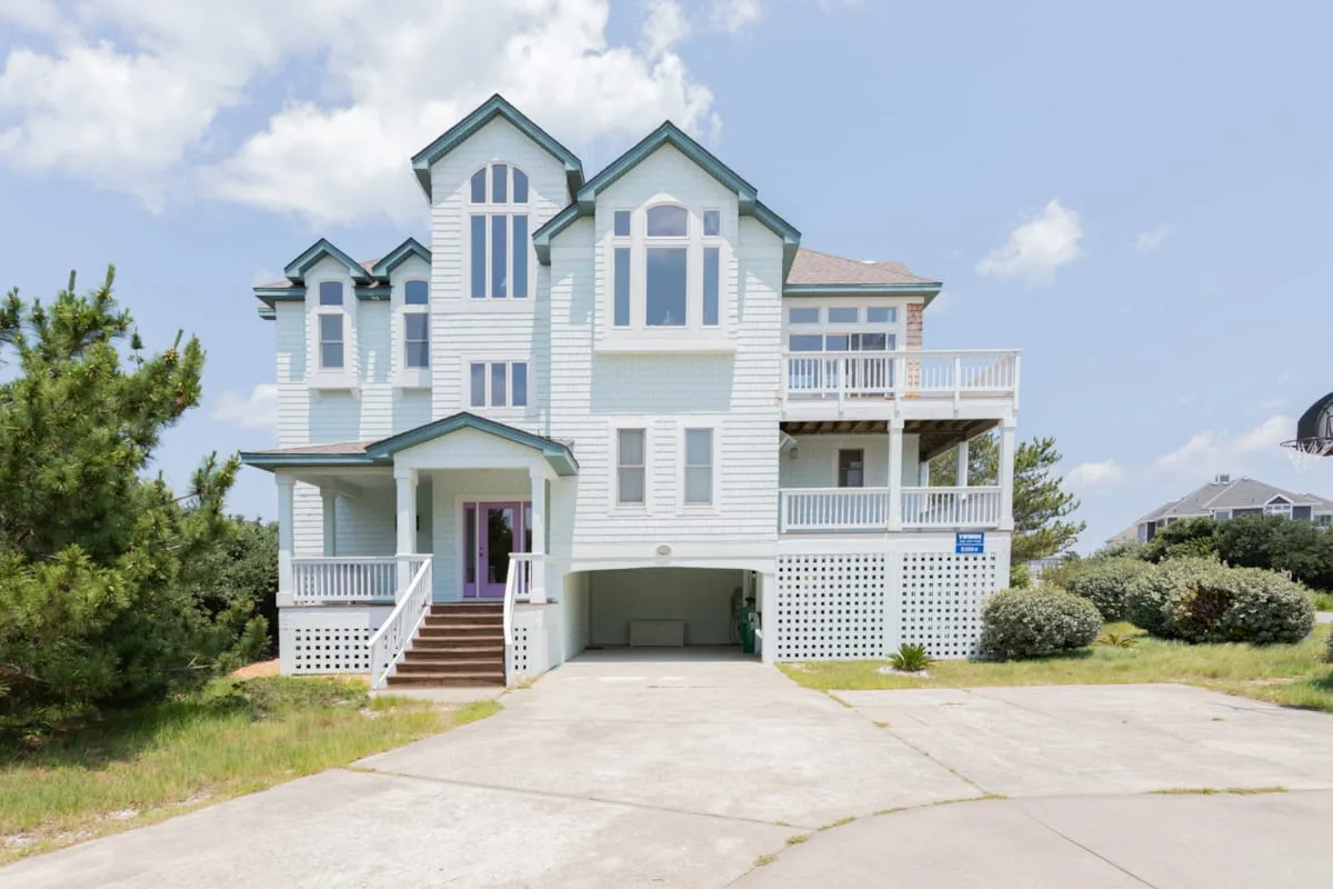A large coastal-style house with white balconies stands beside a paved driveway.