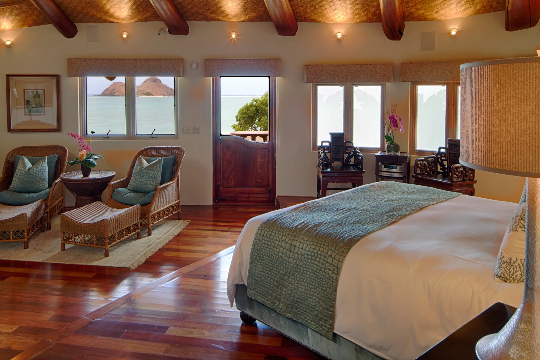 Un dormitorio con una cama grande, asientos de mimbre y ventanas con vistas al agua.